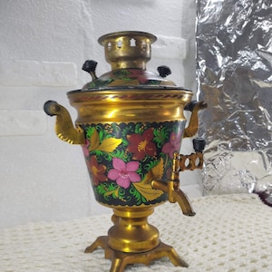 Könnte beinhalten: Ein goldener Samowar mit floralen Malereien in Rot, Rosa, Grün und Schwarz. Der Samowar hat einen Ausguss und einen Griff auf jeder Seite. Er steht auf einer weißen gehäkelten Oberfläche.