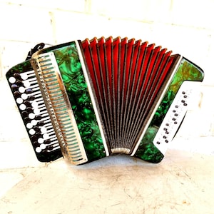 May include: A button accordion featuring a vibrant green marbled finish on its casing. The instrument includes a series of white and black circular buttons on both sides, a metallic grille, and a central bellows section with deep red inner folds.