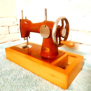 Peut inclure: Une machine à coudre vintage, corps brun rougeâtre et socle en bois clair. Elle comprend un volant, une aiguille et un petit tiroir. Le design rappelle les anciennes technologies de couture.