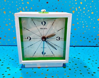 May include: A white square alarm clock with a green trim and a white face. The clock face has black hands, black numbers, and the words "Sevani" and "4 Jewels" printed on it. The clock is made in the USA.