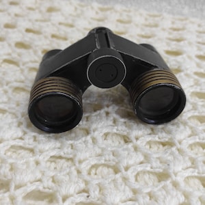 Peut inclure: Jumelles noires avec des accents dorés et une sangle en cuir noir. Les jumelles ont une surface texturée et sont nettes.