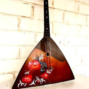 May include: A traditional balalaika, a Russian stringed instrument, with a triangular wooden body. The instrument features a dark brown neck and a body painted with red flowers and a bird design. The balalaika is leaning against a white brick wall.