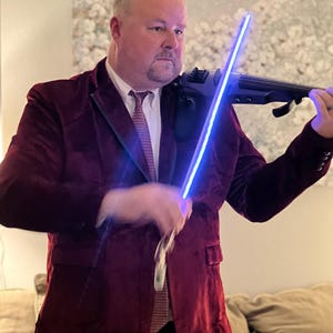 May include: A person wearing a deep red velvet blazer, a white collared shirt, and a patterned tie, playing a black electric violin with a glowing blue LED bow. The instrument features a sleek, modern design with visible electronic components.