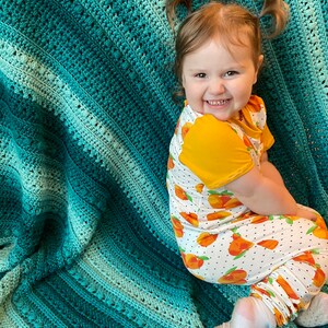 May include: A young child with pigtails smiles while sitting on a teal and green crocheted blanket. The child is wearing a white and yellow outfit with a peach print.