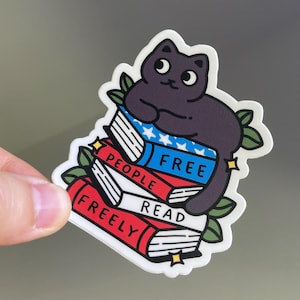May include: A sticker featuring a cartoon black cat sitting on a stack of books. The books are red, white, and blue, with the words "FREE PEOPLE READ FREELY" printed on them. Green leaves and gold stars surround the cat and books.