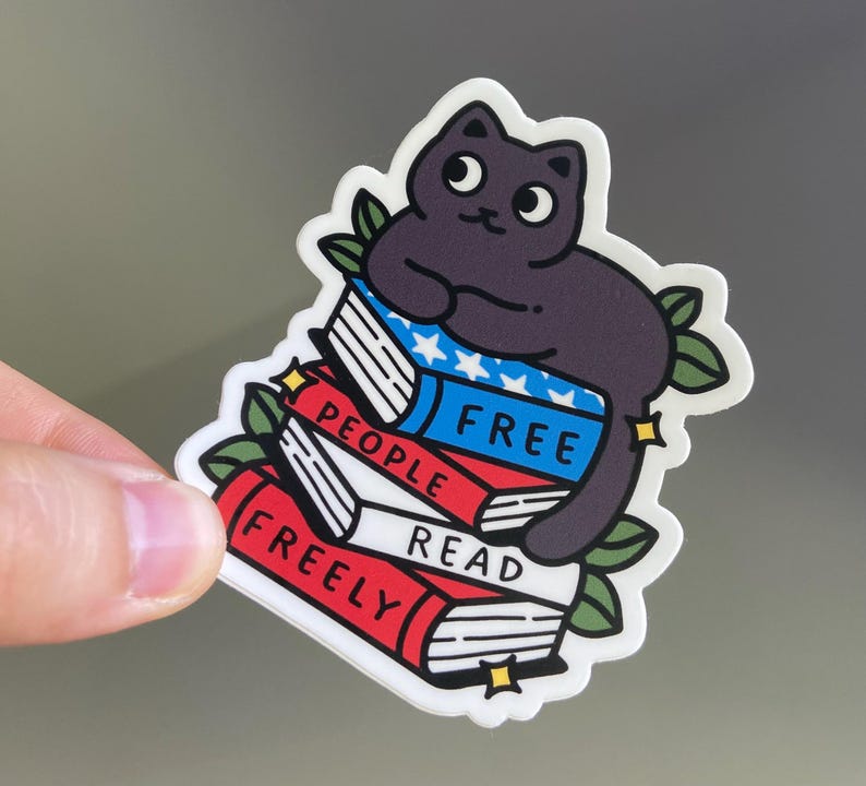 May include: A sticker featuring a cartoon black cat sitting on a stack of books. The books are red, white, and blue, with the words "FREE PEOPLE READ FREELY" printed on them. Green leaves and gold stars surround the cat and books.