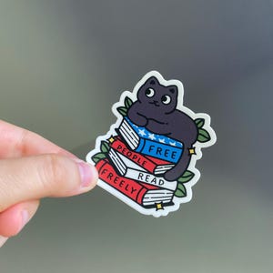 May include: A sticker featuring a black cat sitting atop a stack of books. The books are red, white, and blue, with the words "FREE PEOPLE READ FREELY" printed on them. Green leaves and yellow stars surround the cat and books.