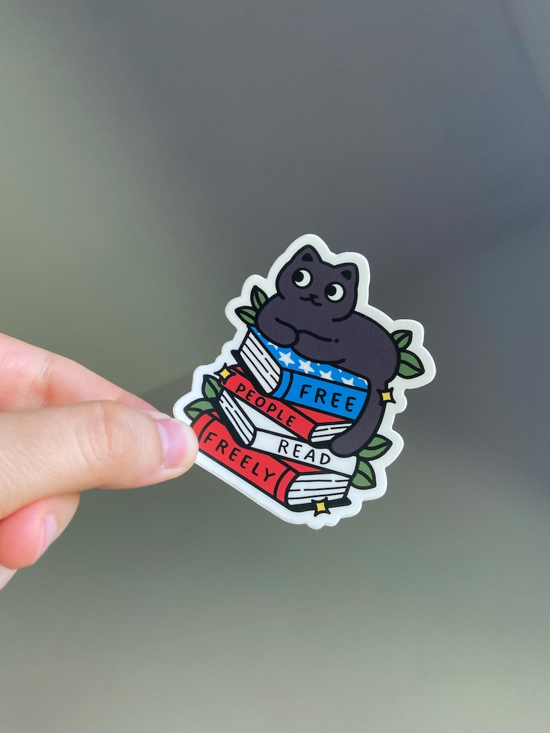 May include: A sticker featuring a black cat sitting atop a stack of books. The books are red, white, and blue, with the words "FREE PEOPLE READ FREELY" printed on them. Green leaves and yellow stars surround the cat and books.