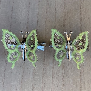 May include: Two green butterfly hair clips with silver accents. The butterflies have a glittery finish and black eyes.