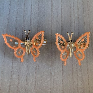 May include: Two gold butterfly hair clips with orange glitter. The butterfly wings are made of a delicate wire mesh.