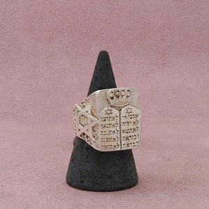 May include: A silver-toned ring featuring a rectangular face engraved with Hebrew text, two small Star of David symbols, and a crown motif at the top. The sides of the ring display a textured, openwork design with a prominent Star of David on each side.