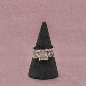 May include: A silver ring featuring three clear, rectangular-cut stones arranged horizontally. The center stone is slightly larger than the two flanking stones, all set in a polished metal band that encircles a dark, textured, cone-shaped display stand.