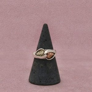 May include: A metal ring featuring two overlapping leaf-shaped elements. One leaf is finished in a silver tone, while the other leaf is finished in a copper tone. The band of the ring is silver-toned and connects to the leaf design at the center.