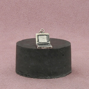 Puede incluir: Un pequeño dije de portátil plateado con un lazo para un collar o pulsera. El portátil está representado con una pantalla y un teclado, y está sobre un objeto cilíndrico gris oscuro. El fondo es de color rosa apagado.