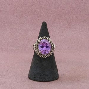 May include: A silver ring featuring a large, oval-cut purple gemstone at the center. The stone is surrounded by a decorative, textured silver border with small, bead-like accents that frame the central purple oval setting.