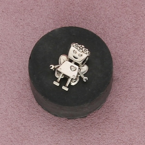 May include: A silver charm in the shape of a robot fairy with wings, a heart on its chest, and a smiling face. The charm is on a dark gray, round surface.