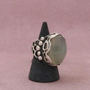 May include: A silver ring featuring a large, smooth, oval-shaped, translucent gray stone set within a textured, openwork metal frame that displays a series of small, raised, circular silver beads along the side of the band.