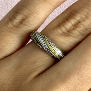 May include: A silver ring with three bands of gemstones. One band is white, one is yellow, and one is silver. The ring is on a finger, with a pink background.