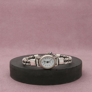 Könnte beinhalten: Eine silberfarbene Armbanduhr mit weißem Zifferblatt und schwarzen Ziffern. Die Uhr hat ein geflochtenes Armband und einen kleinen blauen Edelstein. Die Uhr wird auf einer dunkelgrauen, kreisförmigen Plattform vor rosa Hintergrund präsentiert.