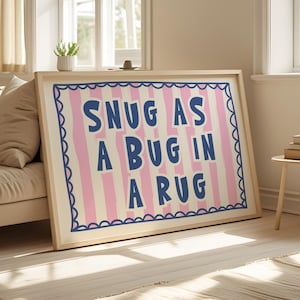 Puede incluir: Un cuadro enmarcado con el texto "SNUG AS A BUG IN A RUG" en azul marino sobre un fondo crema con rayas rosas. El marco es de color madera clara.