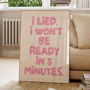 Può includere: Una stampa artistica incorniciata con la scritta "I LIED. I WON'T BE READY IN 5 MINUTES" in rosa. La stampa ha uno sfondo color crema e una cornice dorata. L'opera d'arte è esposta in una stanza ben illuminata con una tavolozza di colori neutri.