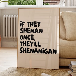 May include: A framed print with the text "If they shenan once, they'll shenanigan" in a bold, black font. The print has a light beige background and a thin, wooden frame. The print is displayed in a living room setting.