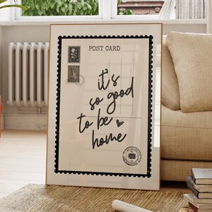 May include: A framed postcard print with the text "it's so good to be home" in black script. The print has a postage stamp border and two stamp illustrations. The frame is gold, and the print is displayed indoors.