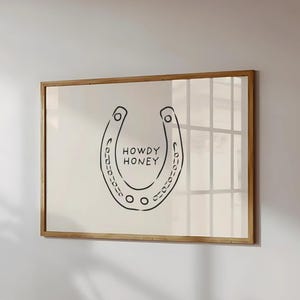 以下が含まれることがあります： 「HOWDY HONEY」の文字と黒い蹄鉄のデザインが特徴のフレーム入りプリント。 作品はクリーム色の背景に配置され、木製のフレームに収められており、壁の装飾に最適です。