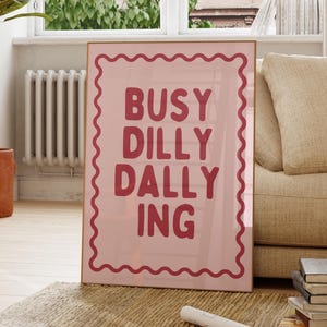 May include: A rectangular wall art print featuring a light pink background with a dark red wavy border. The text BUSY DILLY DALLY ING is printed in the center in a bold, dark red, rounded sans-serif font, arranged in four stacked lines.