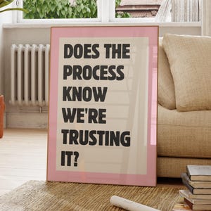 Pode incluir: Um poster emoldurado com uma borda rosa e o texto "DOES THE PROCESS KNOW WE'RE TRUSTING IT?" em preto. O poster é exibido em uma sala bem iluminada com uma janela e um sofá bege.