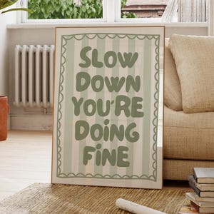 Może przedstawiać: Oprawiony wydruk artystyczny z napisem "SLOW DOWN YOU'RE DOING FINE" w zielonych literach na paskowanym tle. Wydruk ma falisty brzeg i jest eksponowany w pokoju z oknem i sofą.