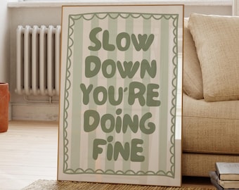 May include: A framed art print with the text "SLOW DOWN YOU'RE DOING FINE" in green letters on a striped background. The print has a scalloped border and is displayed in a room with a window and a sofa.