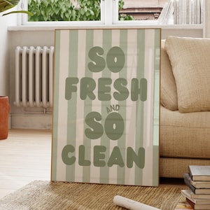 May include: A framed print with a vertical striped background in pale green and off-white. The text "SO FRESH and SO CLEAN" is displayed in large, rounded, pale green letters. The artwork is displayed in a living room setting.