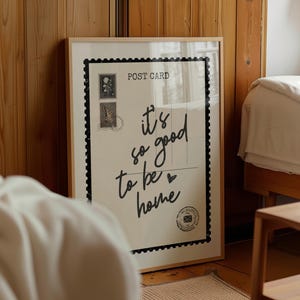 May include: A framed poster with a light wood frame. The poster is designed to look like a postcard with the words "it's so good to be home" in black script. The poster also features two postage stamp illustrations.