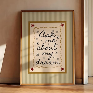 以下が含まれることがあります： 「Ask me about my dream」のテキストが黒い筆記体で書かれた額入りのアートプリント。プリントはクリーム色の背景に装飾的なボーダーと赤い星のアクセントがあります。フレームは温かみのあるゴールド色です。