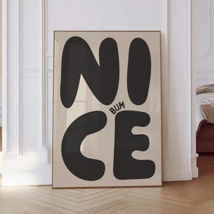 May include: A framed art print featuring the word "NICE" in large, black, rounded letters, with the word "BUM" in smaller letters between the "C" and "E". The background is a light beige colour.