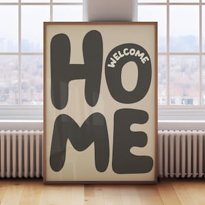以下が含まれることがあります： 大きな濃いグレーの文字で「HOME」と書かれた額入りのアートプリント。文字の「O」には白で「WELCOME」と書かれています。アートワークは、窓と木の床を背景に、明るいベージュ色の背景に表示されています。