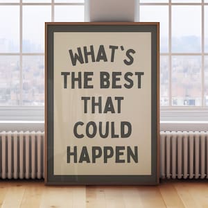May include: Framed poster with the text "WHAT'S THE BEST THAT COULD HAPPEN" in a bold, dark font. The poster has a light beige background and a dark brown frame. The image is set against a window and wooden floor.