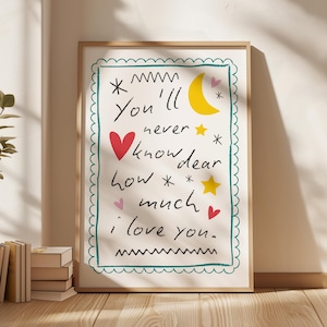 Puede incluir: Una impresión artística enmarcada con el texto "You'll never know dear how much I love you." La obra presenta una luna creciente amarilla, estrellas y corazones en un estilo caprichoso. El marco es de color marrón claro.