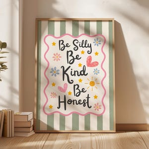 Op de afbeelding: Een ingelijste kunstdruk met de woorden "Be Silly, Be Kind, Be Honest" in zwart schrift. De print heeft een roze golvende rand met sterren, bloemen en harten. De achtergrond is gestreept met groen en wit.