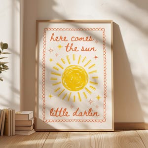 Puede incluir: Un estampado enmarcado con las palabras "here comes the sun little darlin" en escritura naranja. El estampado presenta una ilustración de sol amarillo con rayos y estrellas sobre un fondo crema, enmarcado en un marco de madera clara.