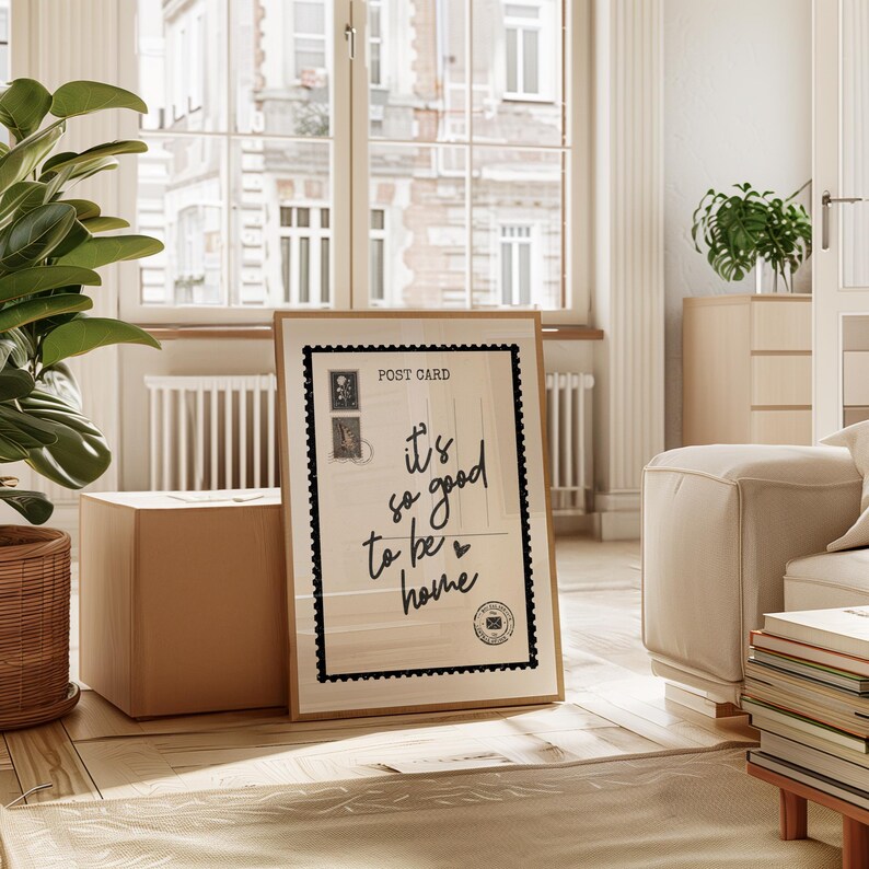 May include: A framed postcard print with the text "it's so good to be home" in black script. The print has a black border and features postage stamp illustrations. The print is displayed on a wooden floor.
