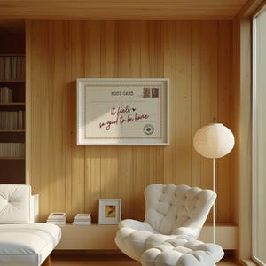 May include: A framed postcard with the text "it feels so good to be home" hangs on a wood-paneled wall. The postcard has a vintage design with postage stamps. A white tufted chair and ottoman are in the foreground.