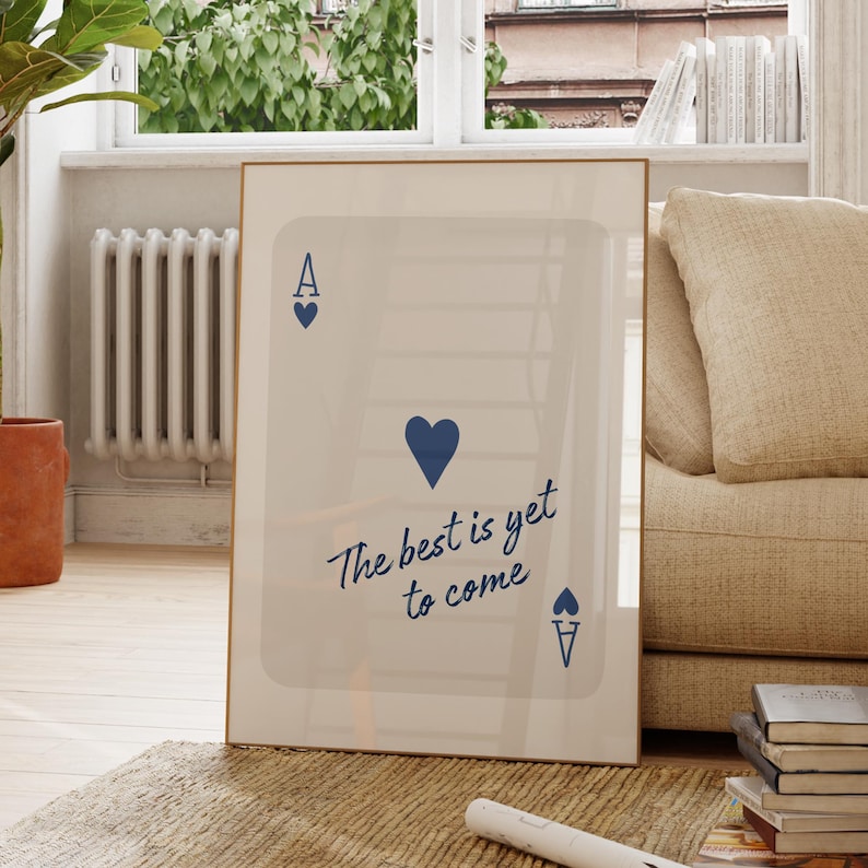 May include: A framed art print featuring a playing card design with the Ace of Hearts. The print has a light background with the text "The best is yet to come" in a handwritten style. The frame is a light brown color.