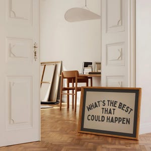 May include: A framed art print with the text "WHAT'S THE BEST THAT COULD HAPPEN" in black lettering. The frame is wood with a black border. The print is displayed on a wooden floor in a room with white walls and a desk.