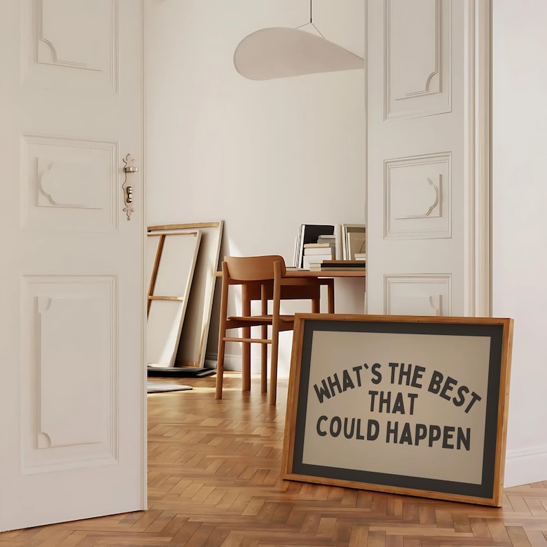 May include: A framed art print with the text "WHAT'S THE BEST THAT COULD HAPPEN" in black lettering. The frame is wood with a black border. The print is displayed on a wooden floor in a room with white walls and a desk.
