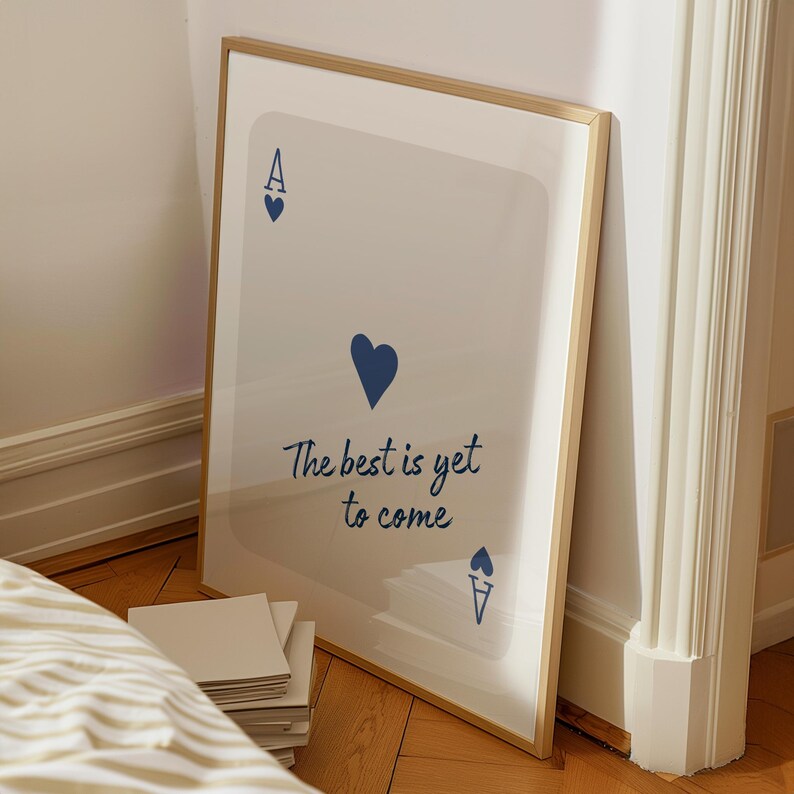 May include: A framed print featuring a playing card design with a blue heart suit and the text "The best is yet to come." The artwork has a light background and a gold-colored frame. The print is leaning against a wall.