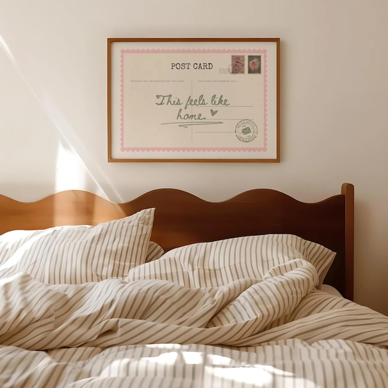 May include: A framed postcard print with the text "This feels like home" hangs above a bed. The postcard has a pink border and decorative stamps. The bed has striped bedding and a wooden headboard.