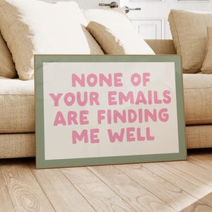 May include: A framed print with the text "NONE OF YOUR EMAILS ARE FINDING ME WELL" in pink letters. The print has a light green border and is leaning against a beige couch. The background includes a wooden floor.