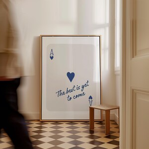 May include: A framed art print featuring a playing card design with an ace of hearts and the text "The best is yet to come" in a stylish font. The print is displayed in a room with a small wooden side table and a checkered floor.